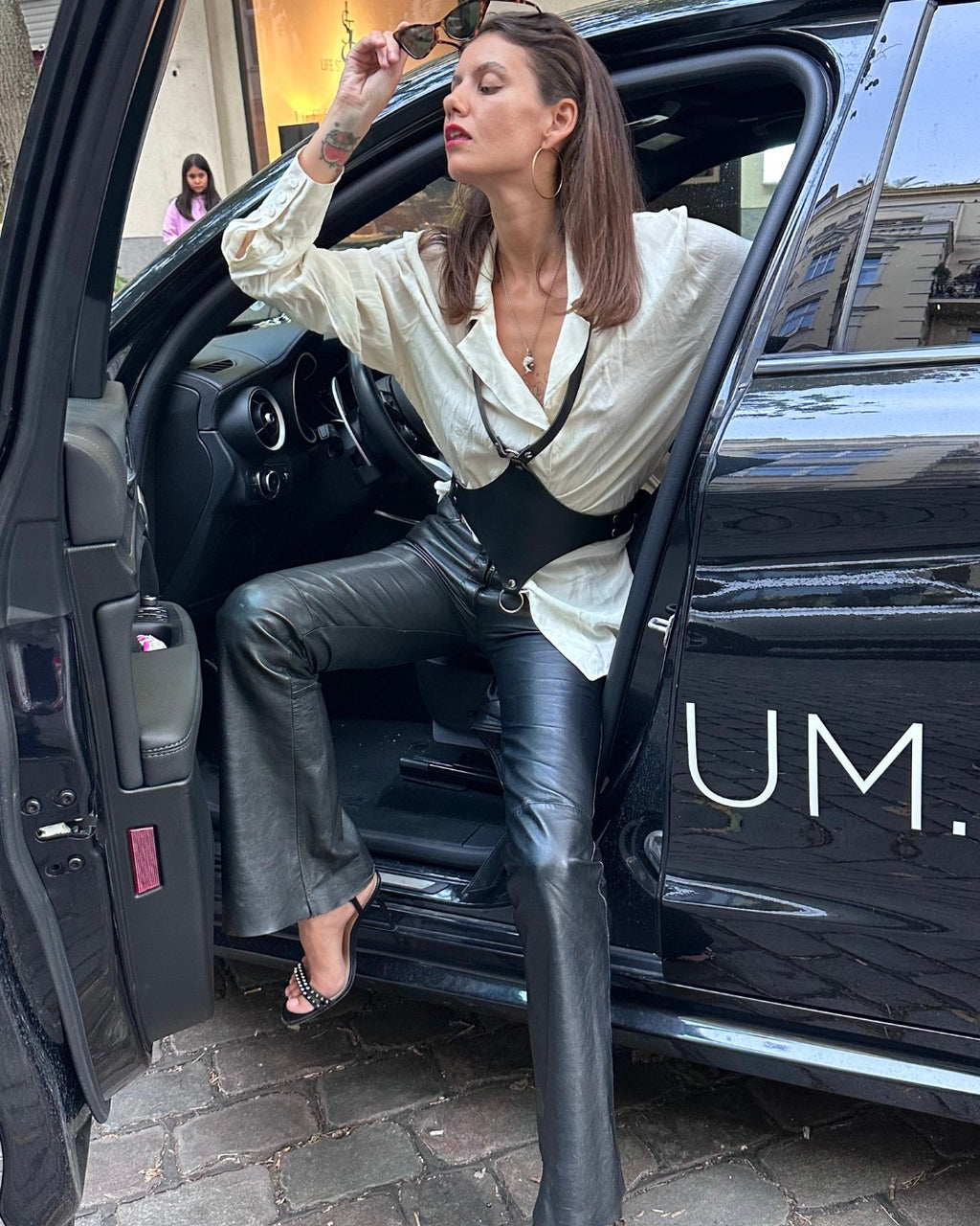 Model trägt schwarzes Lederaccessoire über eleganter Bluse beim Aussteigen aus schwarzem Auto – XMORENIUM-NERO