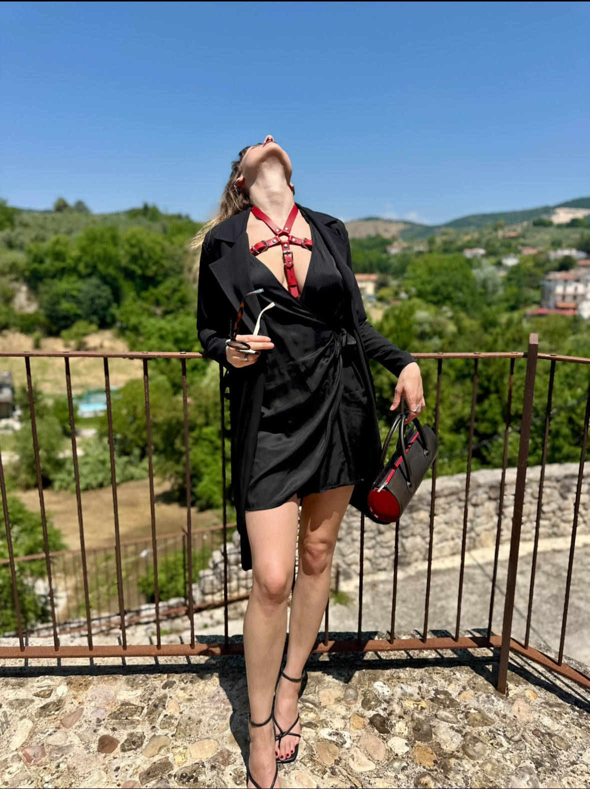 RedTulip Leder Accessoire in Rot  - das Basis Modell, auf der Haut unter einem schwarzen Kleid. Hier blitzt der obere Teil des  RedTulip  auf.  Dazu Absatzschuhe und eine schwarz-rote Handtasche. Ganzkörperaufnahme - XMORENIUM-NERO
